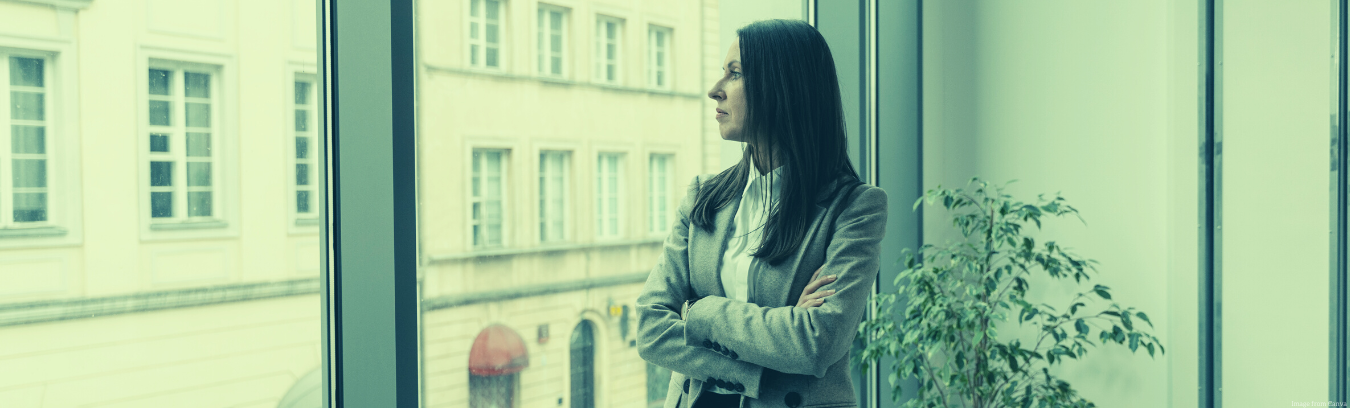 Image of a dark haired woman in a beige blazer and white collared shirt staring out the window towards the street and tan building with a red awning.